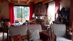 Un salon chaleureux avec un canapé, des fauteuils et une table à manger. Il y a une fenêtre avec des rideaux rouges, un meuble en bois et un vase rempli de fleurs.