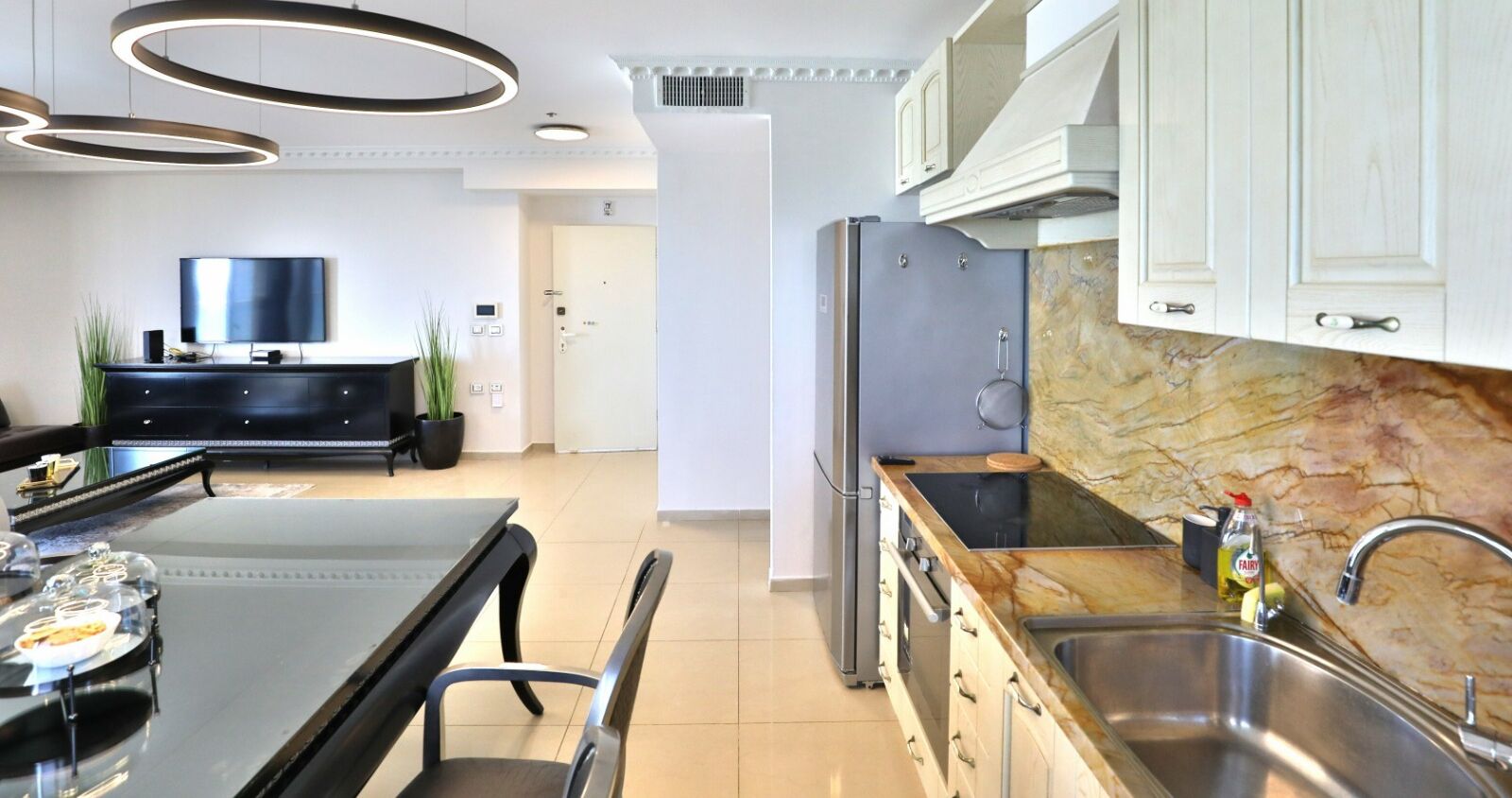 A modern kitchen with a marble countertop, stainless steel sink, and a built-in oven. There is a dining area with a glass table and chairs, and a flat-screen TV in the background.