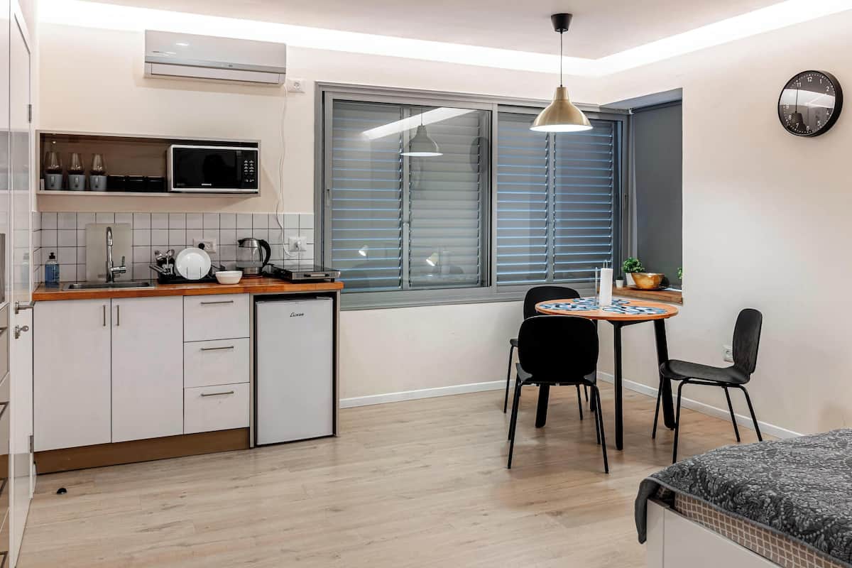 A compact kitchen with a microwave, sink, and a small refrigerator. There is a dining area with a table and chairs, and a window with blinds.