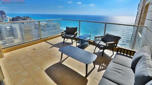 Ein Balkon mit einer Couch, zwei Stühlen und einem kleinen Tisch, mit Blick auf Stadt und Meer.