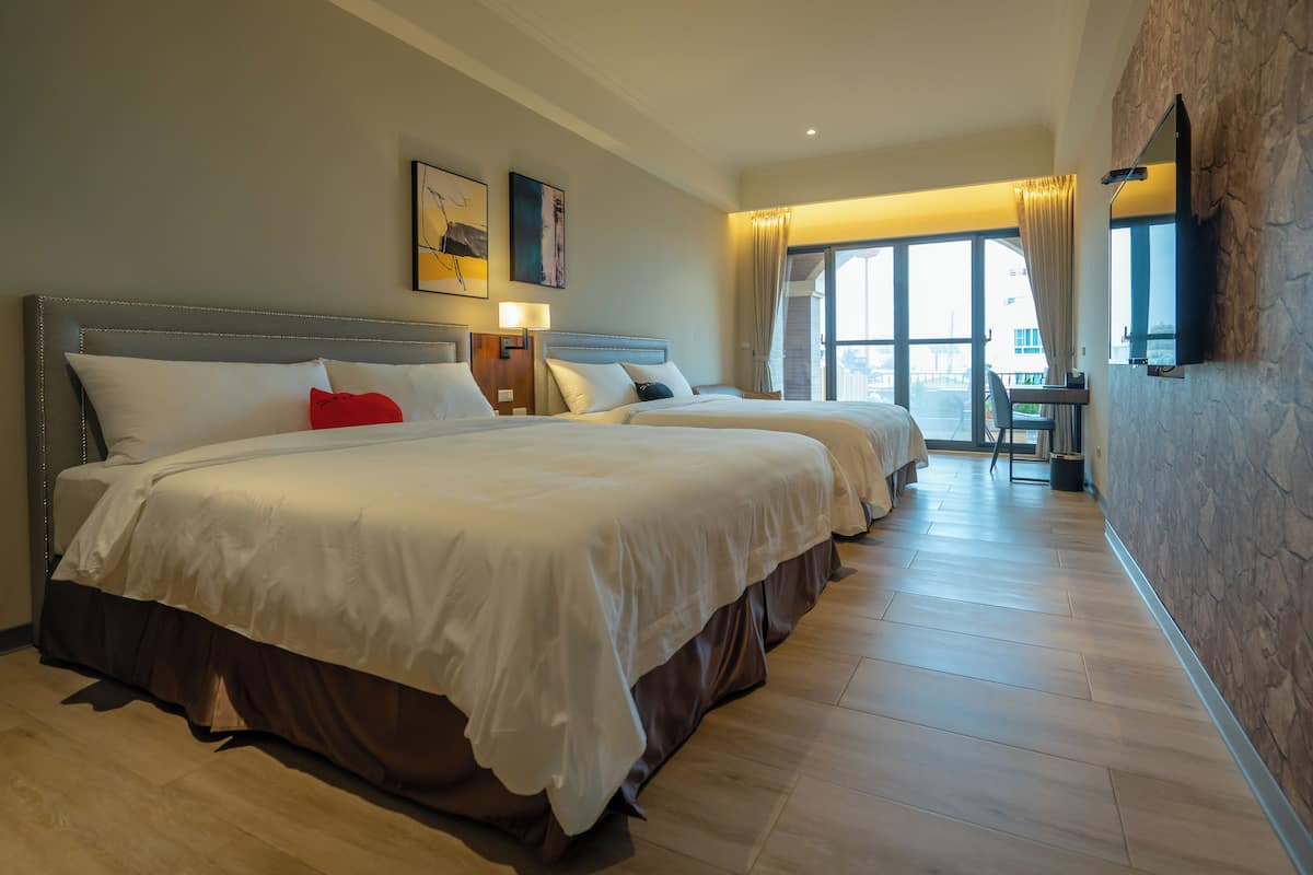 A hotel room with two large beds, a flat-screen TV mounted on the wall, and a large window with curtains.