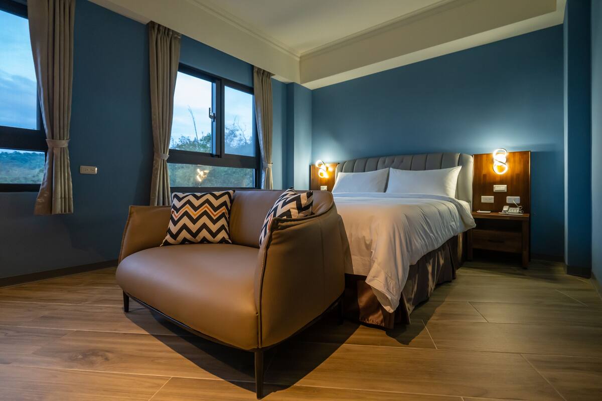 A modern hotel room with a brown leather sofa, a large bed with a grey headboard, and two bedside tables with lamps.
