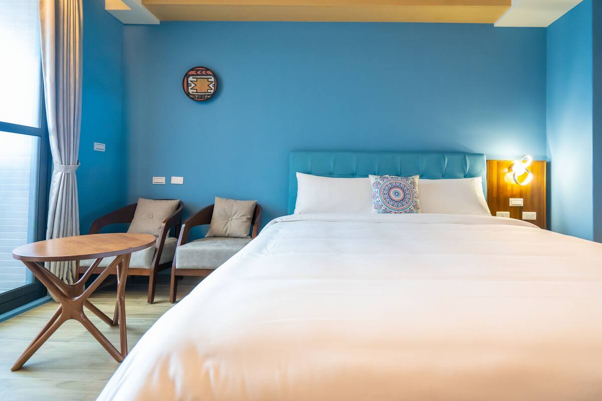 A hotel room with a large bed, a round wooden table, two chairs, a blue wall, and a decorative plate on the wall.