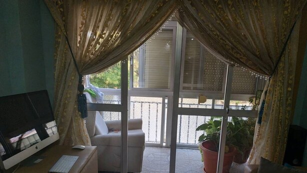 Een kamer met een bureau, een computer, een stoel, een bank en een glazen schuifdeur die leidt naar een balkon met potplanten.