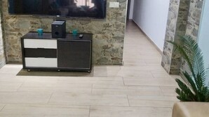 Een woonkamer met een stenen muur, een flatscreen televisie bevestigd boven een zwart-wit dressoir, een gang met ingelijste foto's en een plant.