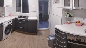 Een moderne keuken met een ingebouwde wasmachine, een fornuis en een spoelbak. Er is een raam met uitzicht op buiten, en een prullenbak is zichtbaar.