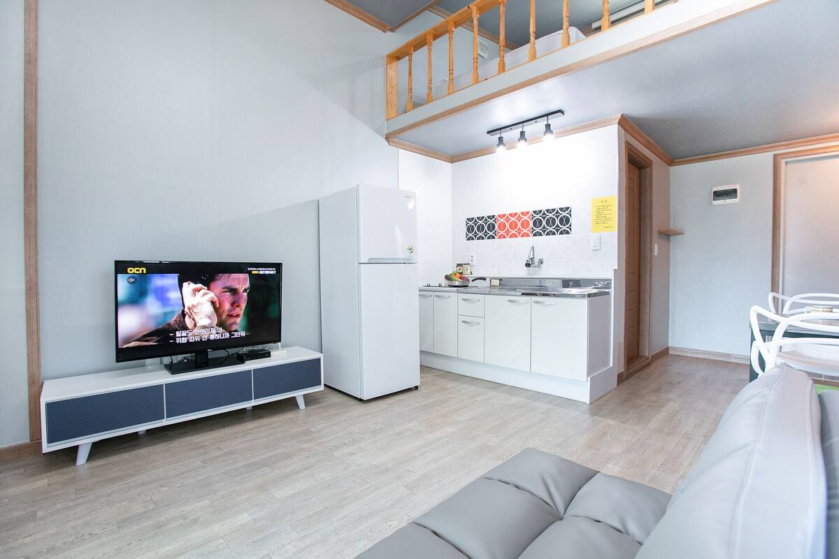 A modern living room with a flat-screen TV, a sofa, a kitchen with white cabinets, and a wooden staircase.