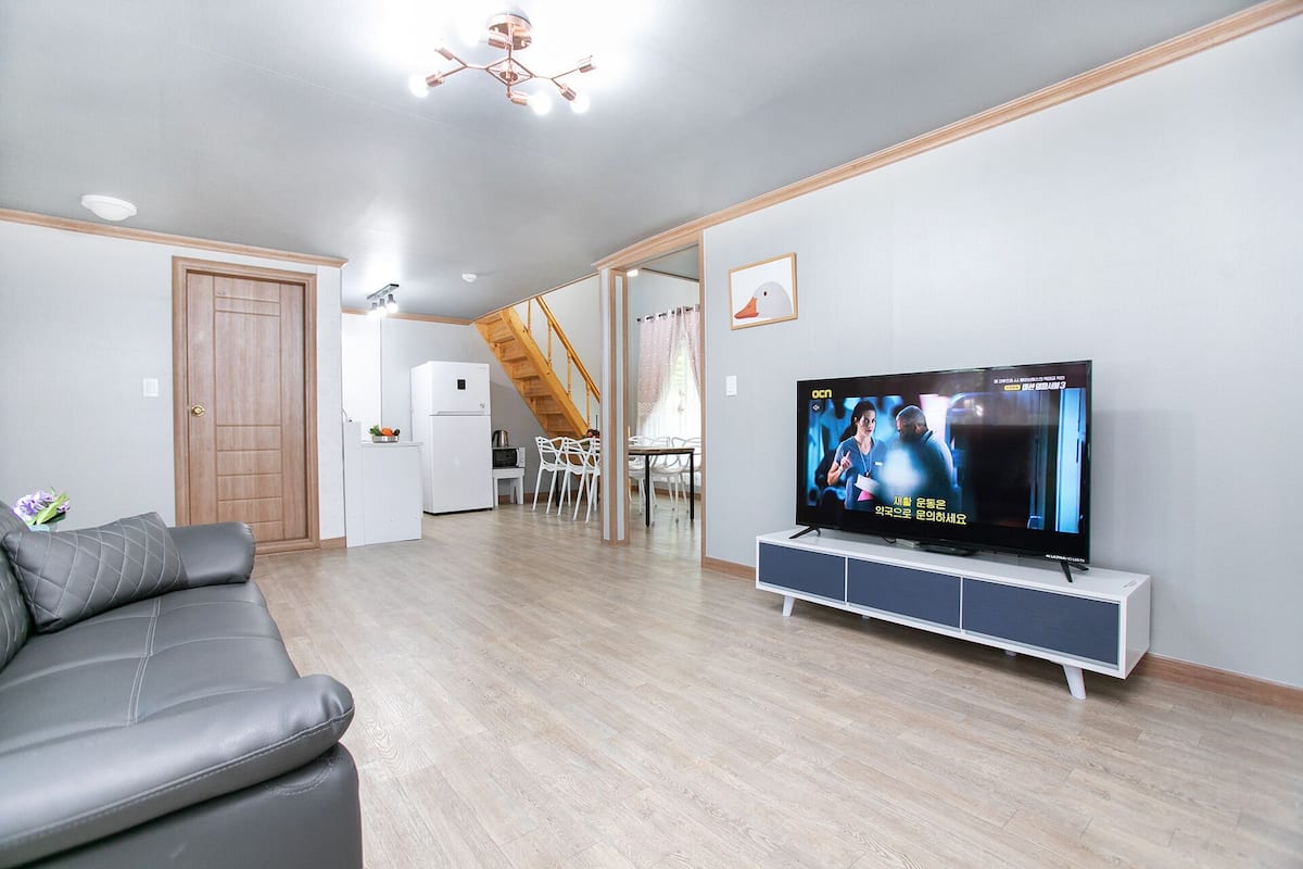 A modern living room with a sofa, a TV on a cabinet, a kitchen area, and a staircase leading to another level.