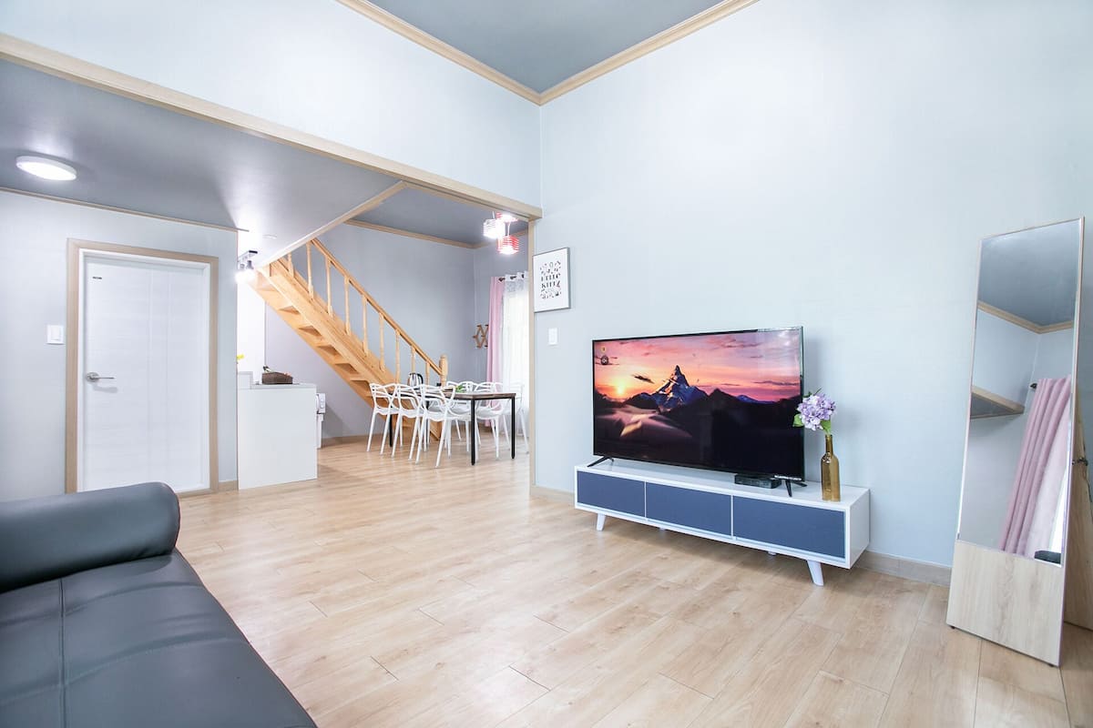 A modern living room with a flat-screen TV, wooden flooring, a grey sofa, a dining area with white chairs, and a staircase.