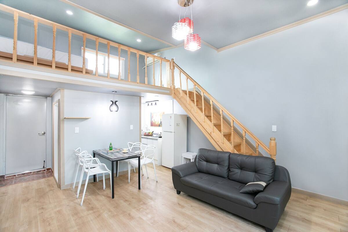 A modern apartment with a loft-style bedroom, a dining area with white chairs, a gray sofa, and a wooden staircase.