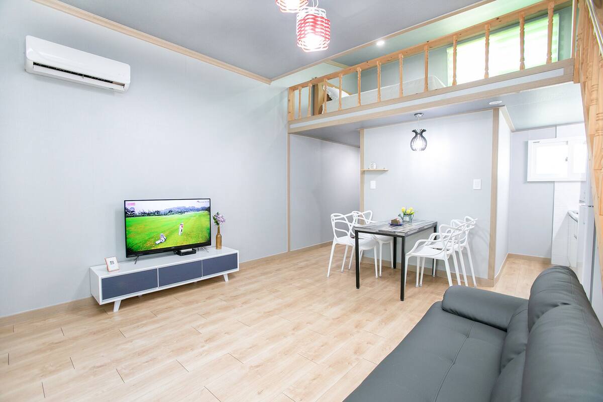 A modern living room with a flat-screen TV, a dining area with white chairs, a grey sofa, and a wooden ceiling.