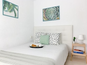 Cama bem arrumada com cabeceira branca, duas almofadas com padrão de bolinhas e uma almofada de destaque verde. Há uma bandeja com croissants e uma xícara na cama. Um pequeno criado-mudo de madeira com abajur e um livro fica ao lado da cama.