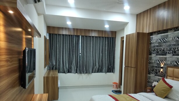 Une chambre d’hôtel moderne avec une armoire en bois, un lit recouvert d’une courtepointe rouge et jaune, un téléviseur fixé au mur et une grande fenêtre avec des rideaux gris.