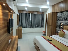 A modern hotel room with a wooden wardrobe, a bed with a red and yellow bedspread, a TV mounted on the wall, and a large window with grey curtains.