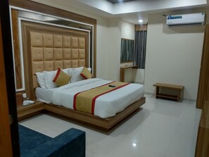 A hotel room with a large bed, a bedside table, a nightstand, a mirror, a small wooden bench, and an air conditioning unit.
