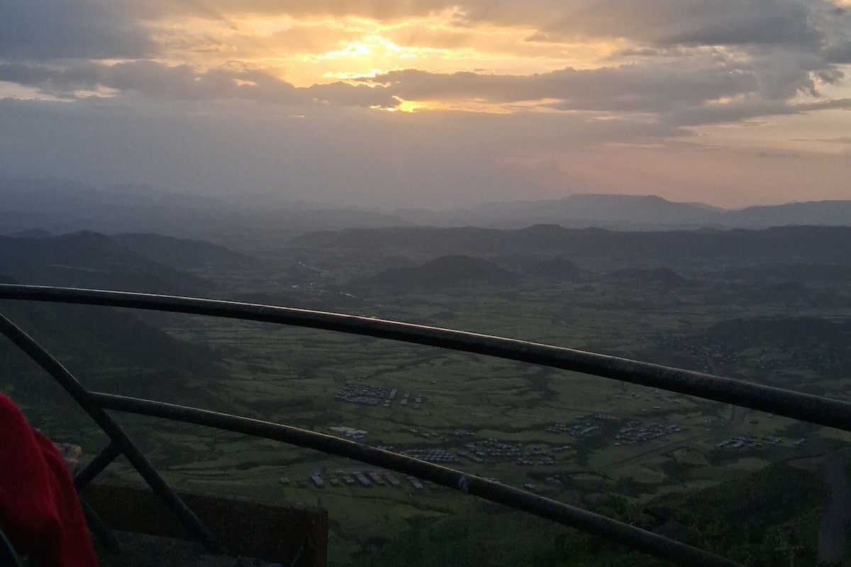A panoramic view from a high vantage point, likely a balcony or terrace, overlooking a valley with distant mountains.