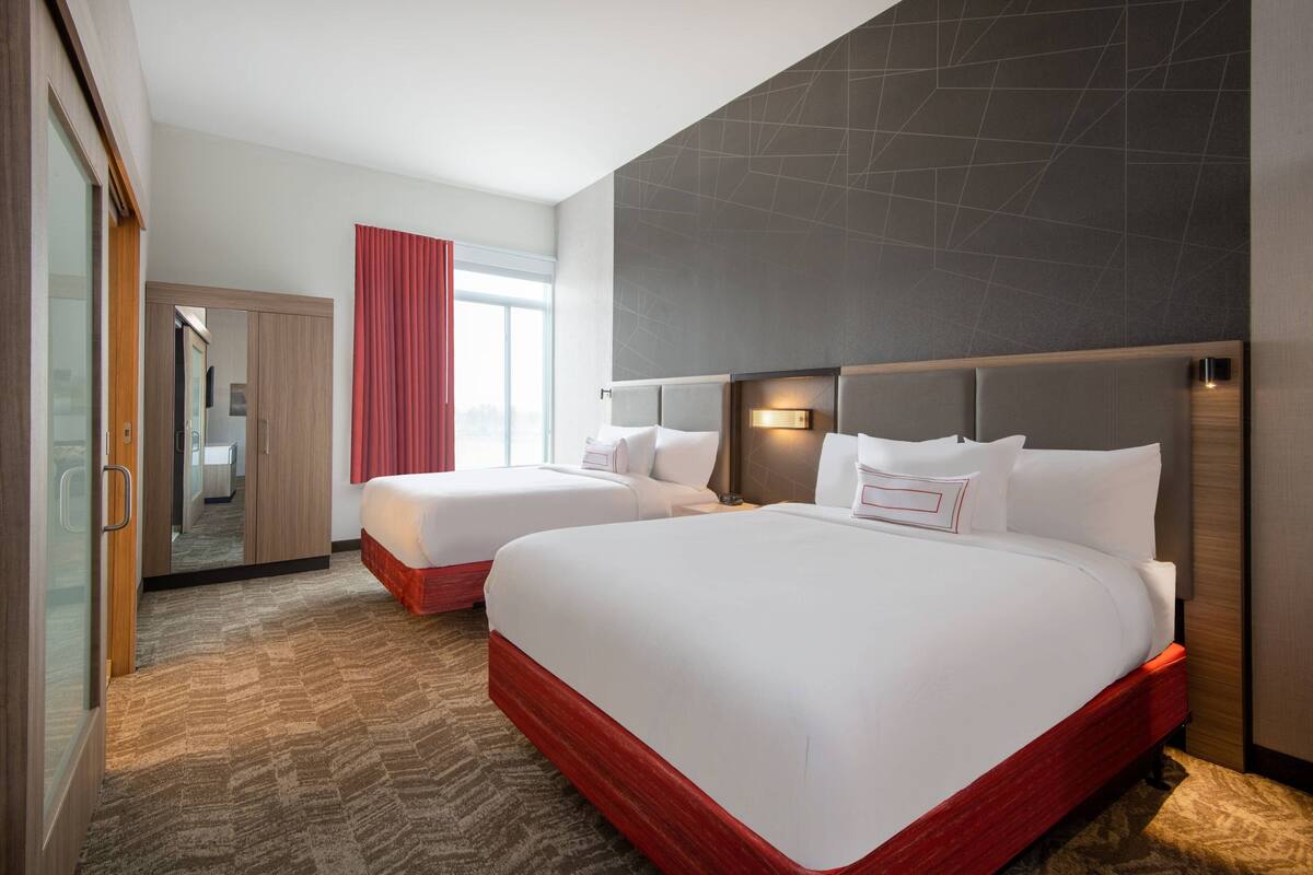 Quarto de hotel com duas camas, um guarda-roupa, um espelho e uma janela com cortinas vermelhas.
