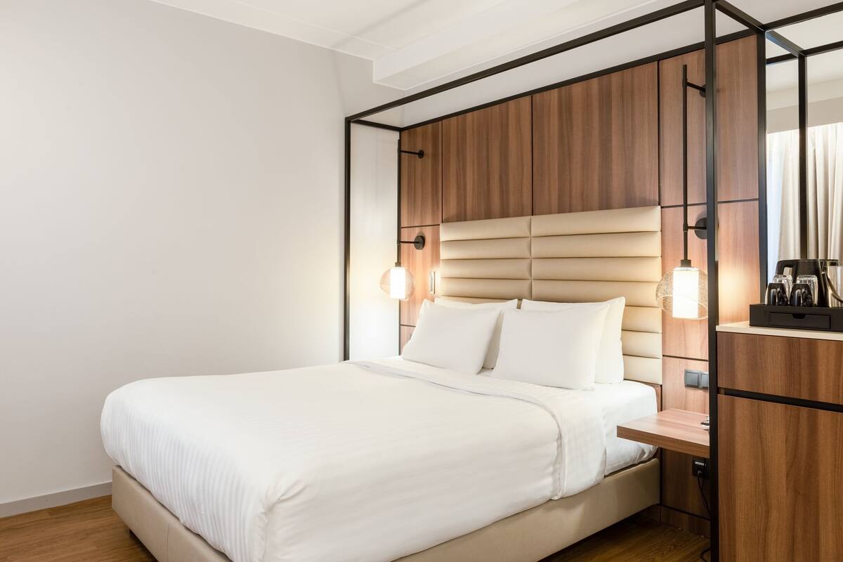 A modern hotel room with a large bed, wooden headboard, bedside tables, and a built-in desk.