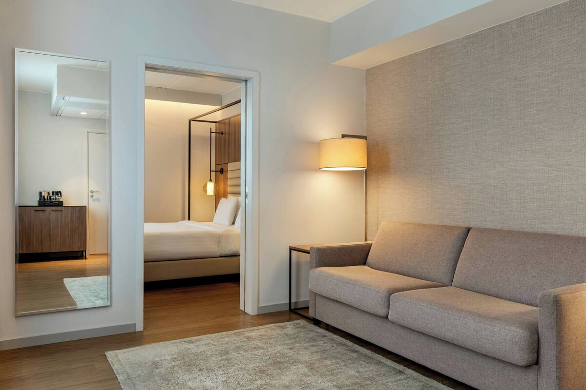 A modern hotel room with a sofa, a bedside table, a lamp, a mirror, and a view into the bedroom.
