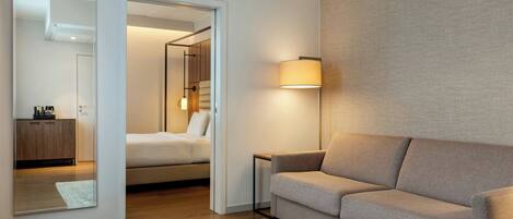 A modern hotel room with a sofa, a bedside table, a lamp, a mirror, and a view into the bedroom.