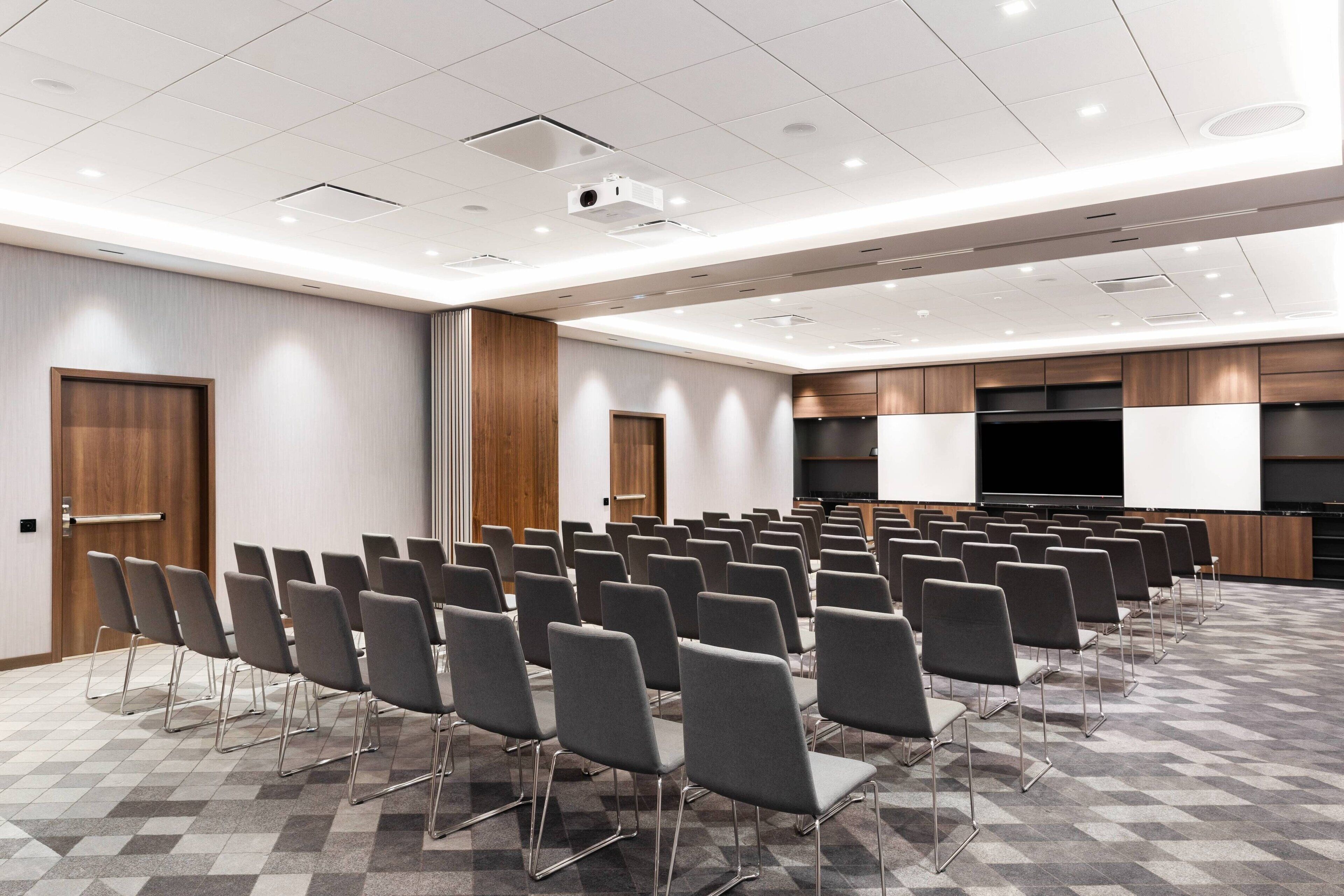 Une salle de conférence avec des rangées de chaises, un grand écran et un mur recouvert de panneaux en bois.