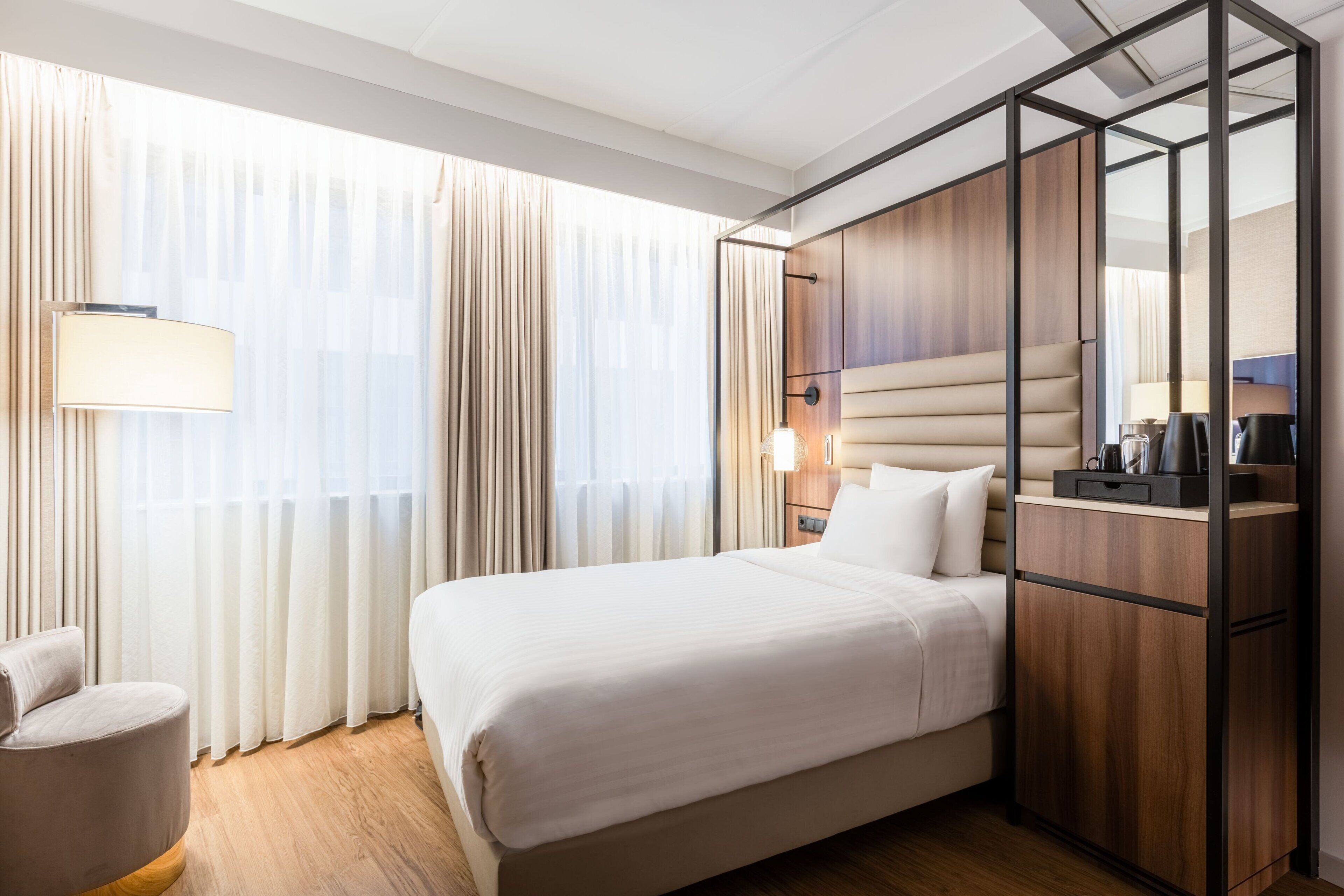 A modern hotel room with a large bed, a wooden wardrobe, a desk with a lamp, and a chair.