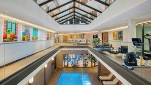 Un atrium moderne dans un hôtel, avec un plafond en verre, une piscine, des équipements de remise en forme et des œuvres d’art colorées qui ornent les murs.