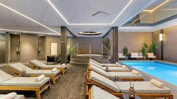 Un área de piscina interior moderna con sillones de descanso, un jacuzzi y una escalera que conduce a un nivel superior.