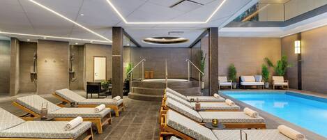 Un área de piscina interior moderna con sillones de descanso, un jacuzzi y una escalera que conduce a un nivel superior.