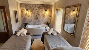 Een spa-ruimte met een groot bad, twee massagetafels en een wand met een bladmotief.