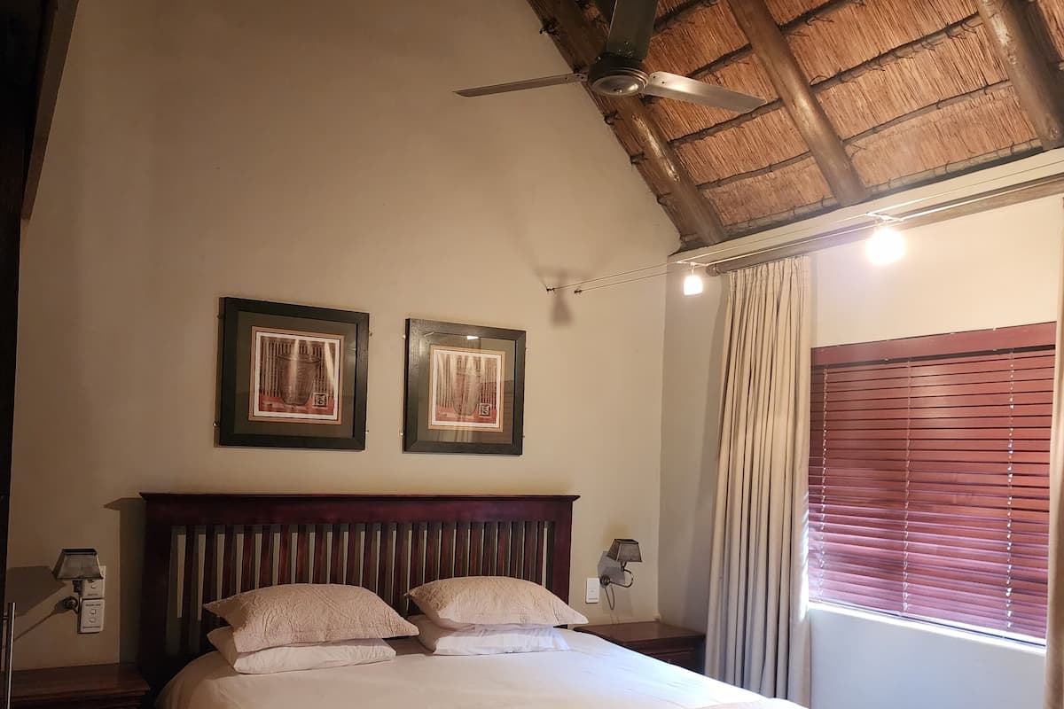 A bedroom with a bed, a wooden headboard, a ceiling fan, two framed pictures on the wall, a nightstand, a lamp, a window with curtains, and a patterned rug.