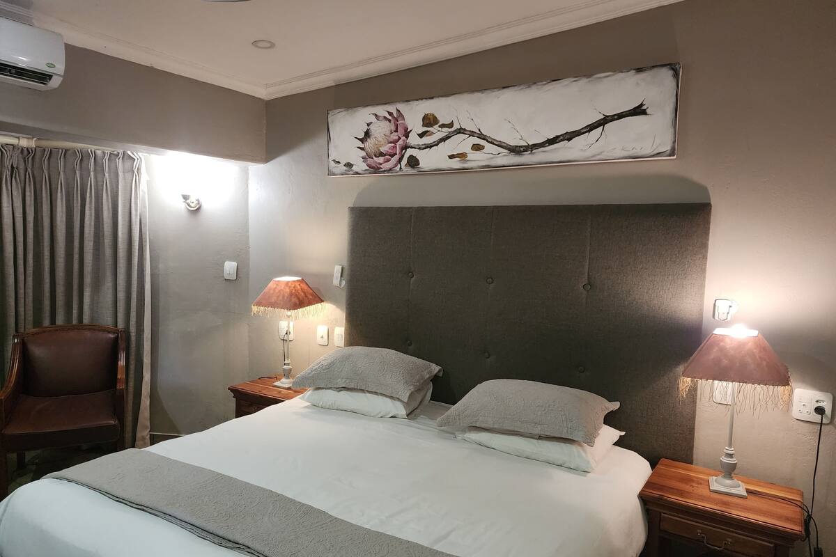 A neatly made bed with a grey and white duvet, two bedside lamps, a brown leather chair, and a wall-mounted artwork.