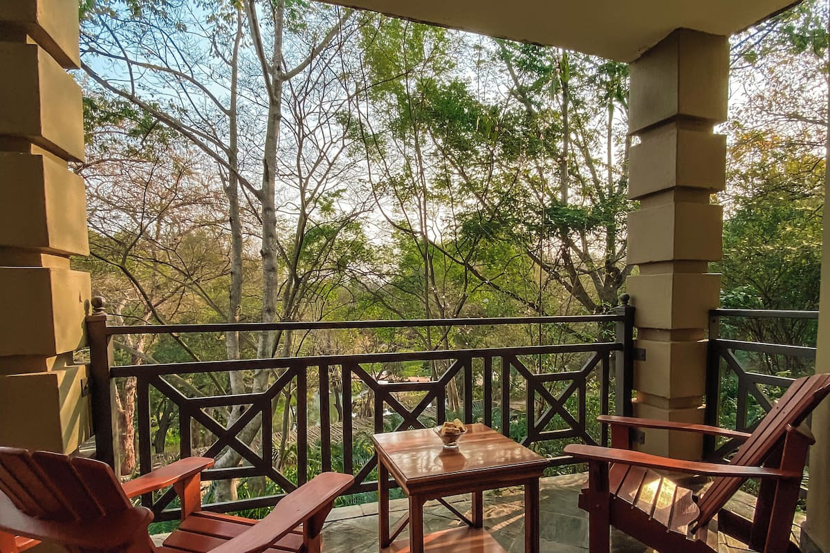 A covered outdoor seating area with two wooden chairs and a small table, surrounded by a railing and overlooking a garden with trees.