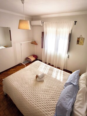 Een slaapkamer met een bed, een hanglamp, een airconditioner, een raam met gordijnen, een radiator en een kleine wanddecoratie.