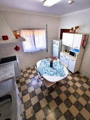 Een compacte keuken met een geruit vloertje, een witte tafel met een blauw geruit tafelkleed, een magnetron, een koelkast en een raam met dunne gordijnen.