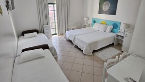 Habitación de hotel con dos camas individuales, un escritorio, una silla, una mesita pequeña, una mesita de noche, una lámpara, un cuadro y una ventana con cortinas.