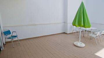 A small, enclosed patio area with a tiled floor, a green umbrella, a blue chair, and a white table with chairs.