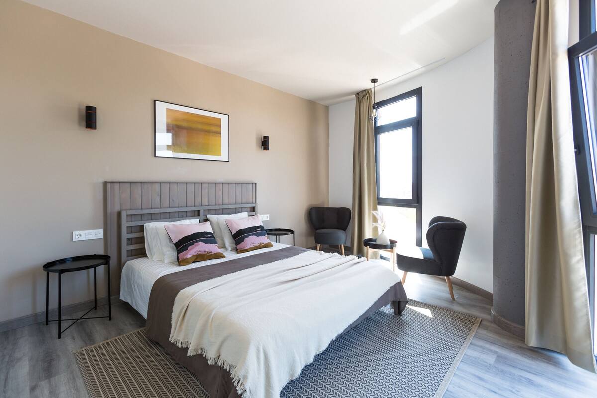 Une chambre d’hôtel moderne avec un grand lit, une petite table de chevet, une chaise et une fenêtre avec des rideaux.