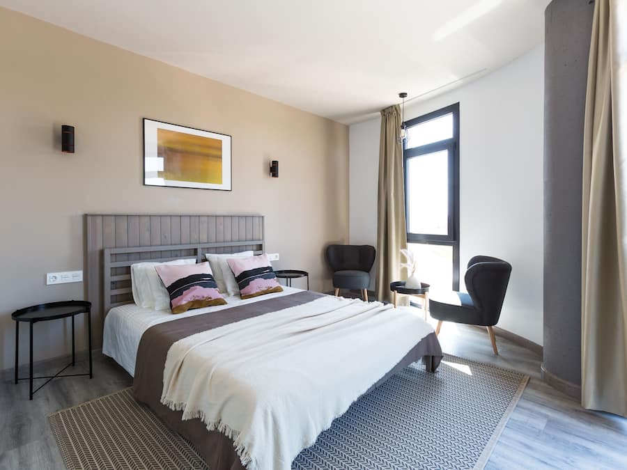 Una habitación de hotel moderna con una cama grande, una mesita auxiliar, una silla y una ventana con cortinas.