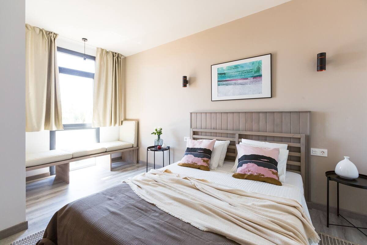 Une chambre à coucher avec un lit, un banc près de la fenêtre, une petite table avec une plante et un tableau accroché au mur.