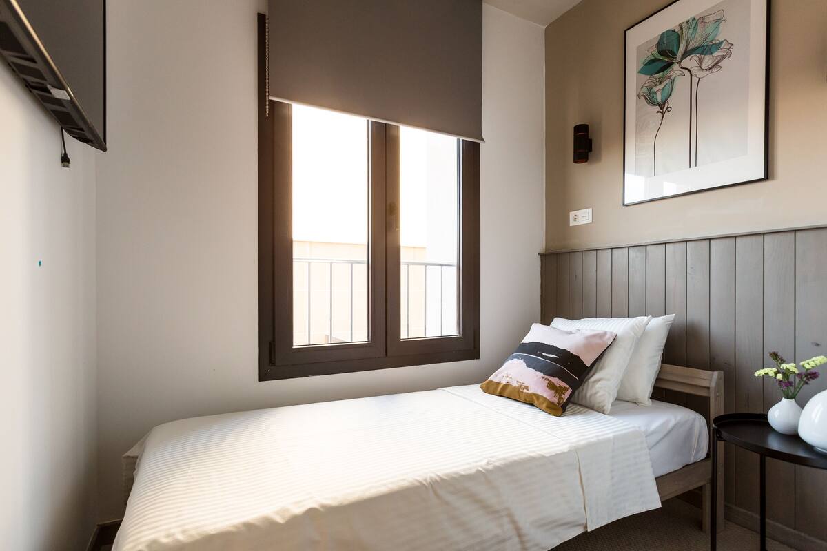 Une chambre à coucher avec un lit, une fenêtre, une œuvre d’art accrochée au mur et une table de chevet sur laquelle se trouve un vase.