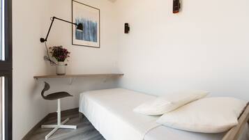 A minimalist bedroom with a bed, a desk with a chair, a wall-mounted lamp, and a framed picture.