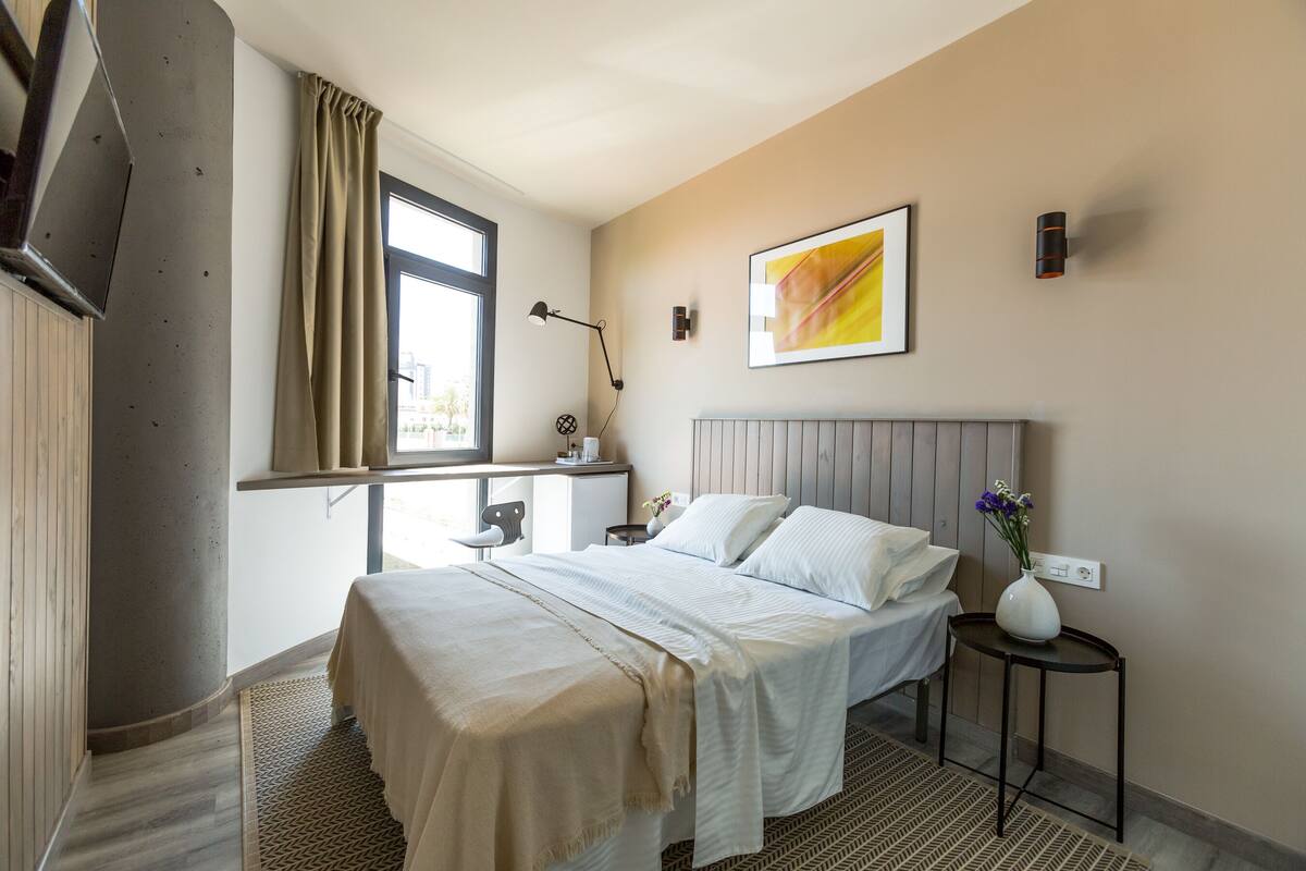 Une chambre d’hôtel moderne équipée d’un lit, d’un bureau, d’une chaise, d’une fenêtre avec des rideaux, d’une lampe fixée au mur et d’un tableau encadré.