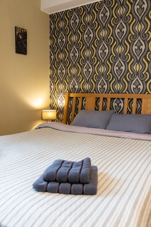 A neatly made bed with a striped bedspread, a wooden headboard, and a patterned wallpaper in shades of gray, yellow, and black.