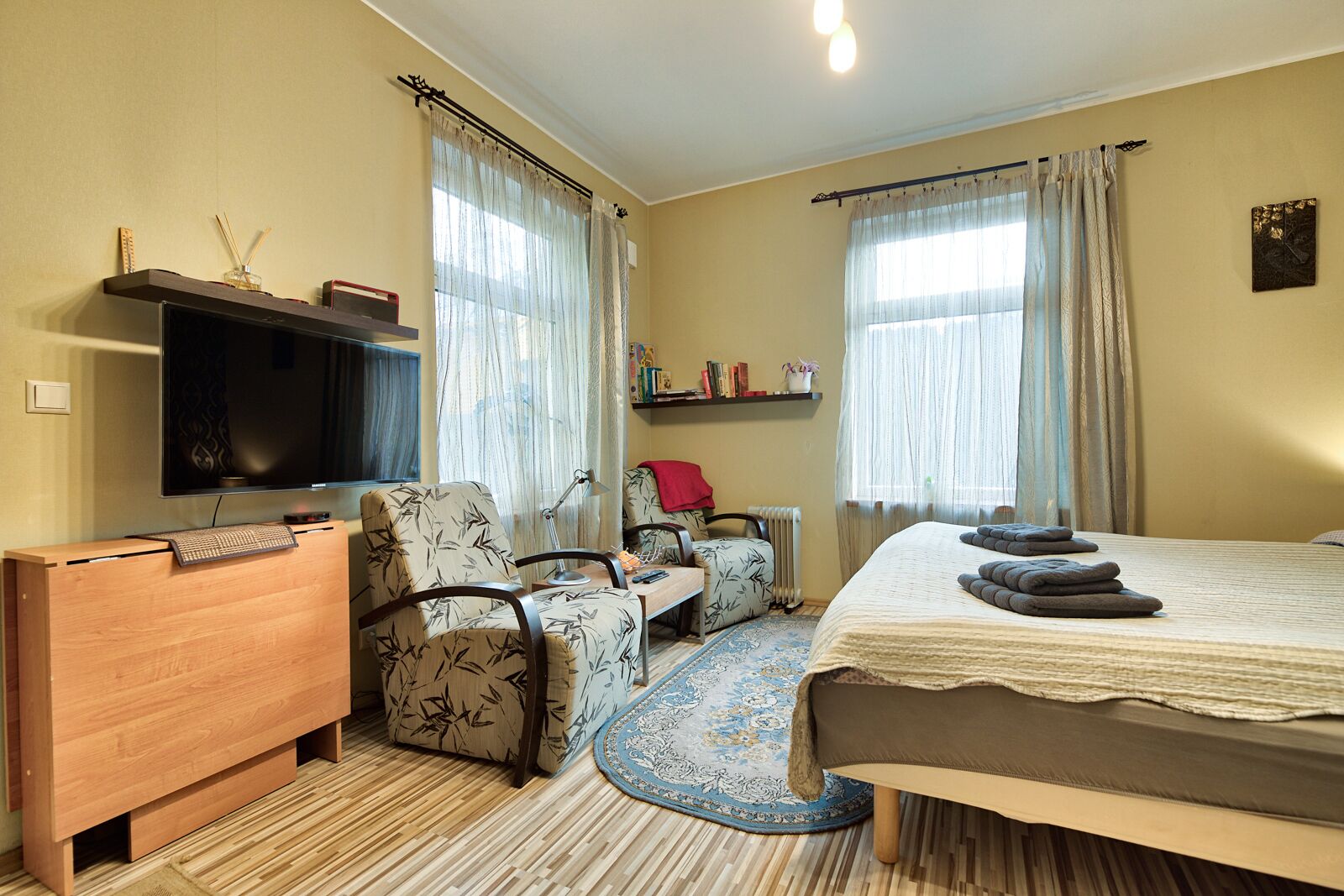 A hotel room with a bed, a desk with a computer, a chair, a TV, and a bookshelf.