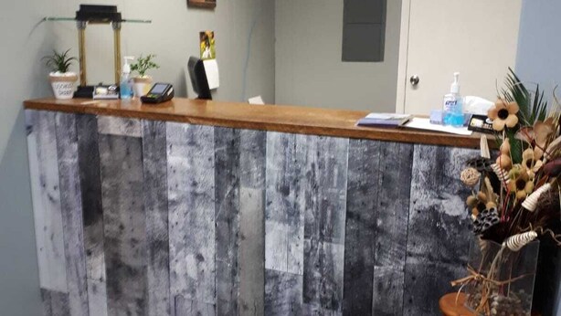 Un comptoir d’accueil avec un plateau en bois et un panneau gris à l’aspect vieilli. On y trouve une petite plante, une chaise et un vase rempli de fleurs séchées, posé sur une table d’appoint.