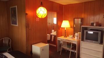 Una habitación de hotel pequeña con paneles de madera, un escritorio con lámpara, un televisor, un refrigerador pequeño y una silla.