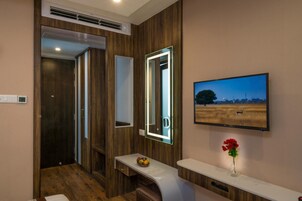 Un baño moderno con paneles de madera, un espejo con iluminación integrada, un televisor en la pared y una pequeña mesa con frutero y un jarrón.
