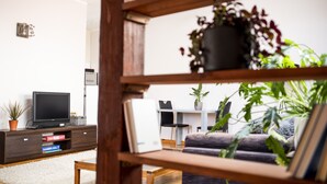 Una sala de estar moderna con un televisor de pantalla plana sobre un mueble de madera, un sofá y una mesa de comedor con sillas. Hay varias plantas en macetas y una pared blanca con un adorno.
