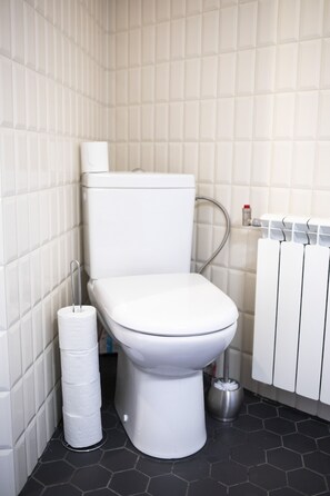 Un baño con inodoro blanco, porta papel higiénico, radiador y un cubo de basura.
